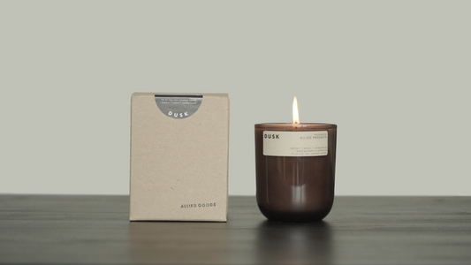 Dusk Soy Wax candle lit in dark glass container next to its cardboard kraft paper packaging. Dusk candle with soothing fragrance and minimalist design, perfect for evening relaxation. Allied Goods Dusk candle with calming scent for a peaceful home ambiance. Luxury candle with clean-burning wax and natural fragrance by Allied Goods. Eco-friendly candle with sustainable ingredients for a refreshing home scent.